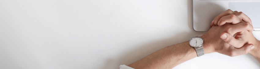 A close-up view of a person's crossed hands resting on a white surface next to a laptop. The person is wearing a silver mesh watch with a white face on their left wrist.