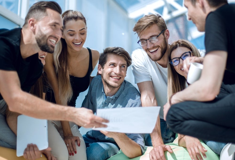 A group of smiling young adults gathered in a semicircle, looking at a sheet of paper that one man is holding and showing to the others. They appear to be engaged in a collaborative discussion or brainstorming session in a bright, modern indoor setting.