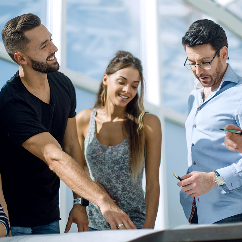 A group of smiling young adults gathered in a semicircle, looking at a sheet of paper that one man is holding and showing to the others. They appear to be engaged in a collaborative discussion or brainstorming session in a bright, modern indoor setting.