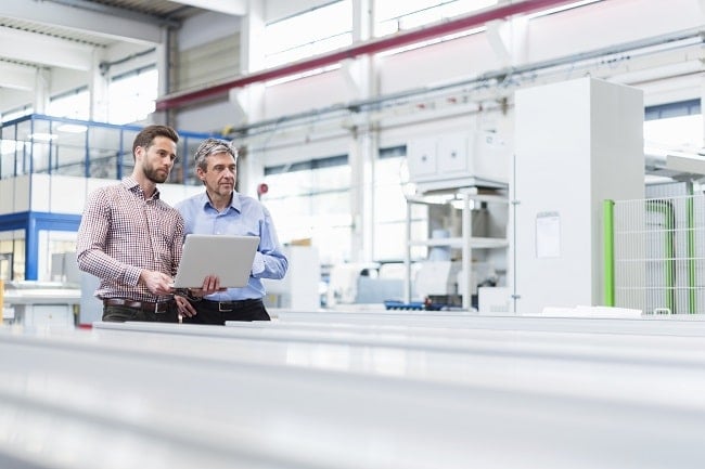Two men standing in a modern industrial facility, looking at a laptop. One is younger and dressed in a checkered shirt, while the older man wears a light blue shirt. They appear to be discussing something related to production or technical operations, with machinery and large equipment in the background.