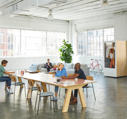 Modern open office space with several people working and conversing. A long wooden table is in the center with chairs around it. Two people are having a discussion at the table, one person is using a laptop at the far end, and another is seated on a couch in the background. A woman is seen standing inside a phone booth-style private pod. The room has large industrial-style windows, polished concrete floors, and a bright, airy atmosphere.