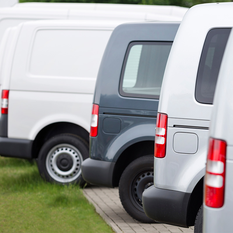 A row of commercial cargo vans parked in a lot, all aligned uniformly. The image shows the rear side of the vehicles, emphasizing their similar design and the consistency of a fleet, likely used for delivery or service operations.
