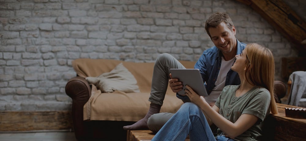 A smiling couple sits together in a cozy living room with exposed brick walls. The woman is holding a tablet while the man looks at it, both appearing engaged and happy. A beige couch with pillows is in the background.