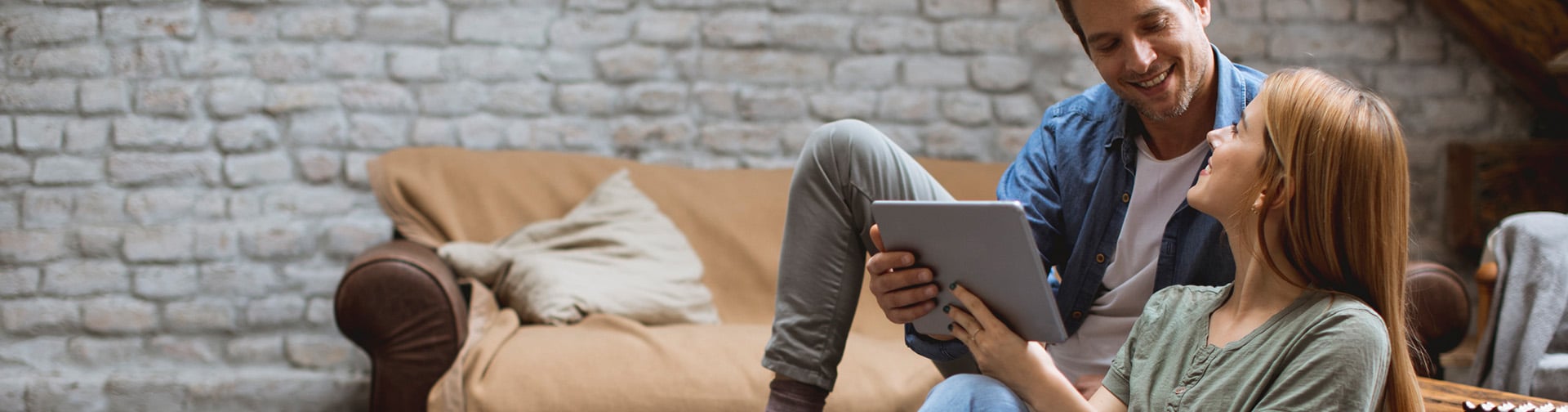 A smiling couple sits together in a cozy living room with exposed brick walls. The woman is holding a tablet while the man looks at it, both appearing engaged and happy. A beige couch with pillows is in the background.