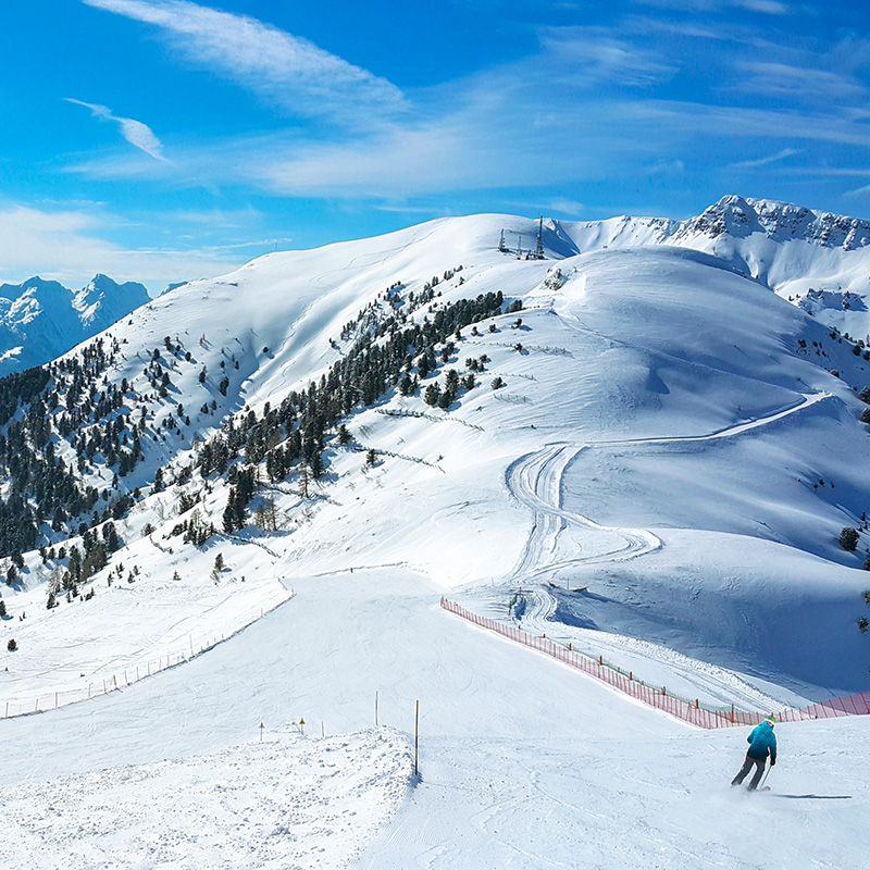 A snowy ski slope with mountains in the background; in the corner, a skier in motion.