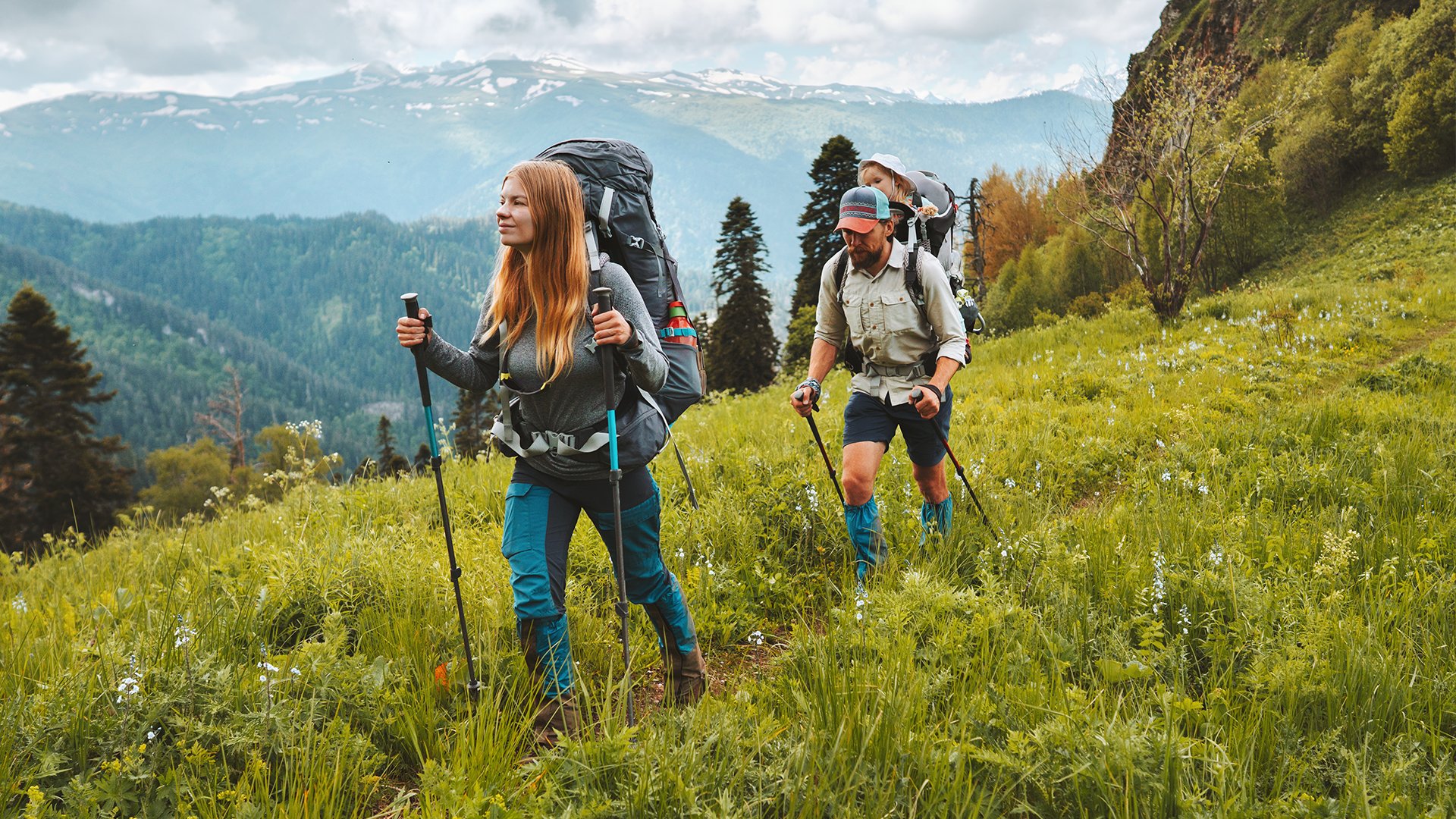 Žena a muž kráčajú po turistickom chodníku uprostred hôr. Obaja sú oblečení turisticky, v rukách majú nordic walking palice. Žena má chrbte ruksak a muž nosič, v ktorom je ich malé dieťa. 