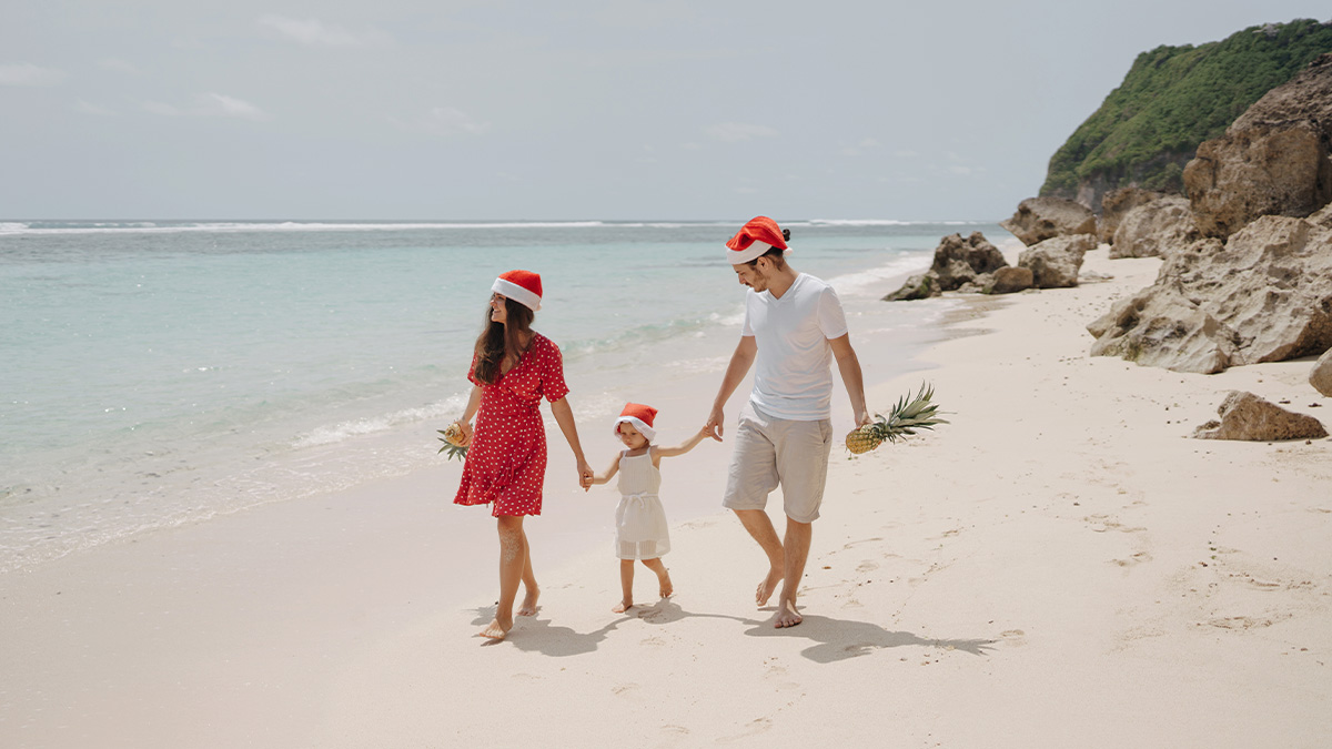 Rodičia s malou dcérou slávia Vianoce v tropickej krajine na pláži. Na hlavách majú mikulášske čiapky.