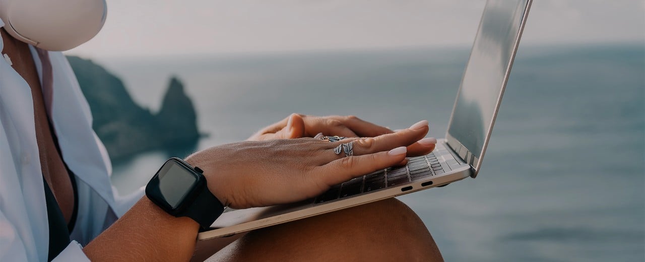 Digitálna nomádka pracujúca na notebooku pri mori. Detail na ruky.