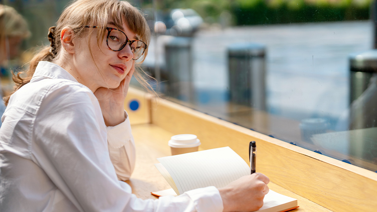 Mladá žena v okuliaroch plánujúca si svoj deň do zápisníka. Nachádza sa v exteriéri a tvári sa zamyslene.