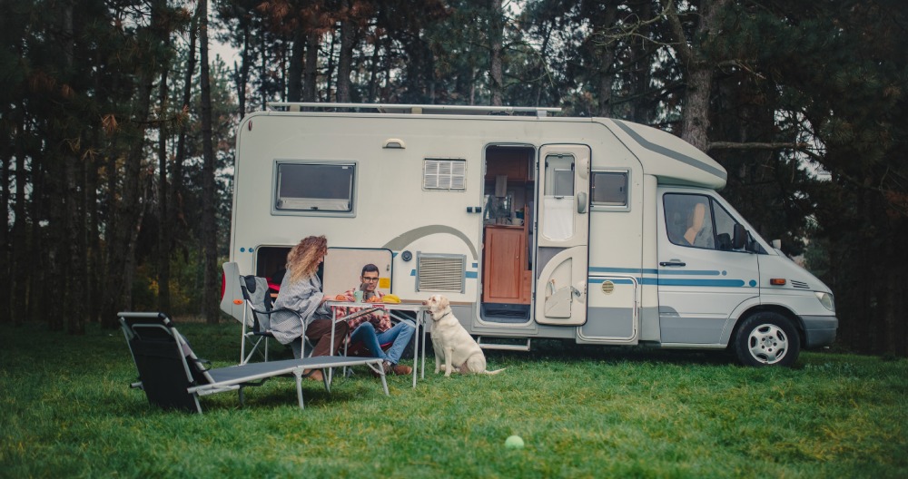 2 osoby so psom sedia v prírode pri karavane.