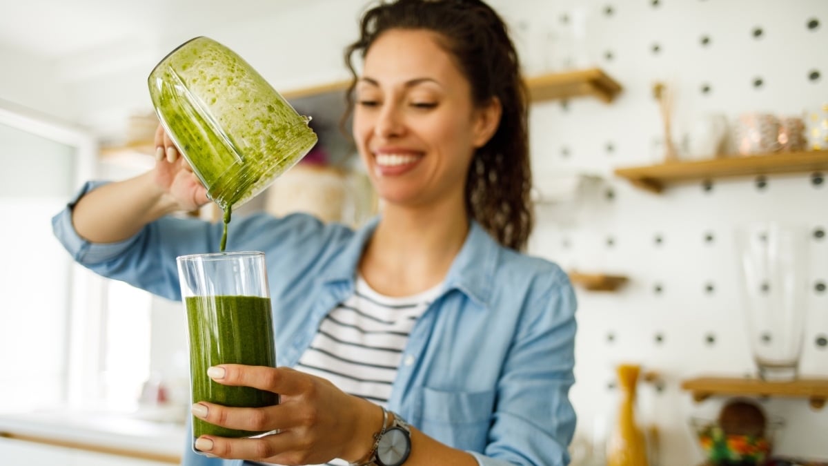 Šťastná usmiata žena v kuchyni prelieva zelené smoothie z mixéra do pohára.