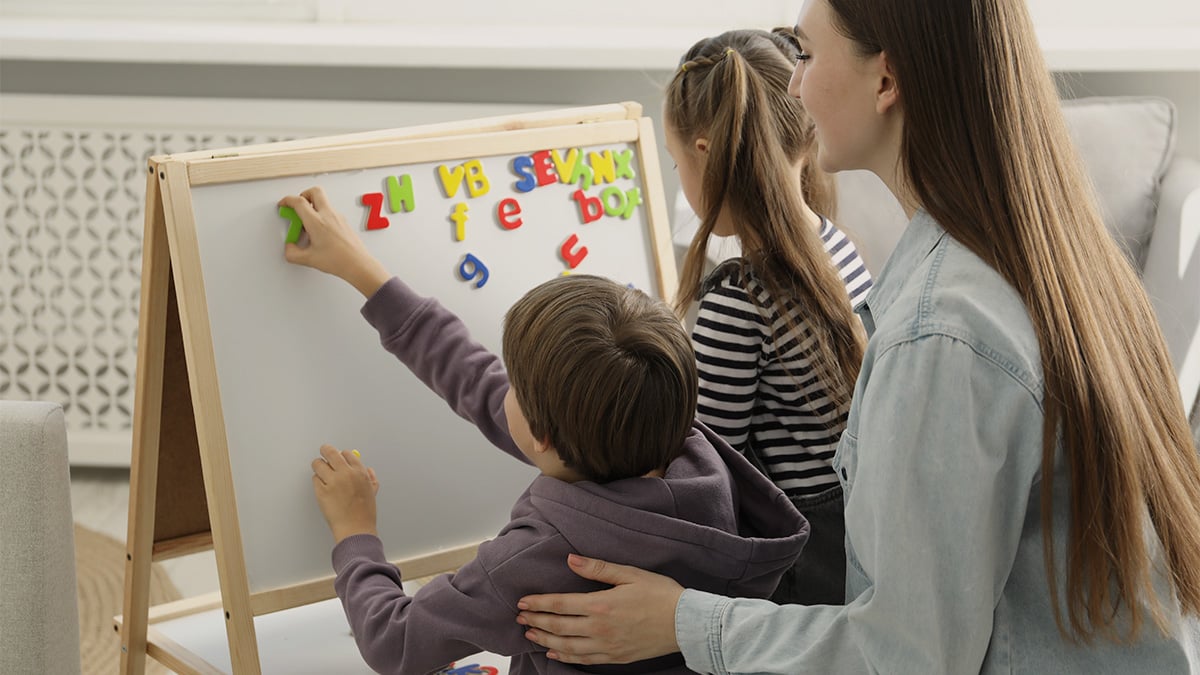 Mladá mama pomáha deťom usporiadať farebné písmenká na bielej magnetickej tabuli.