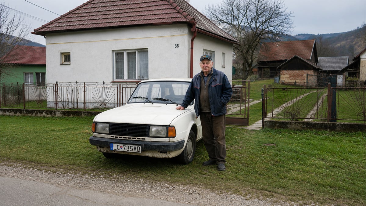 Muž v dôchodkovom veku stojaci pri svojom starom aute pred domom.