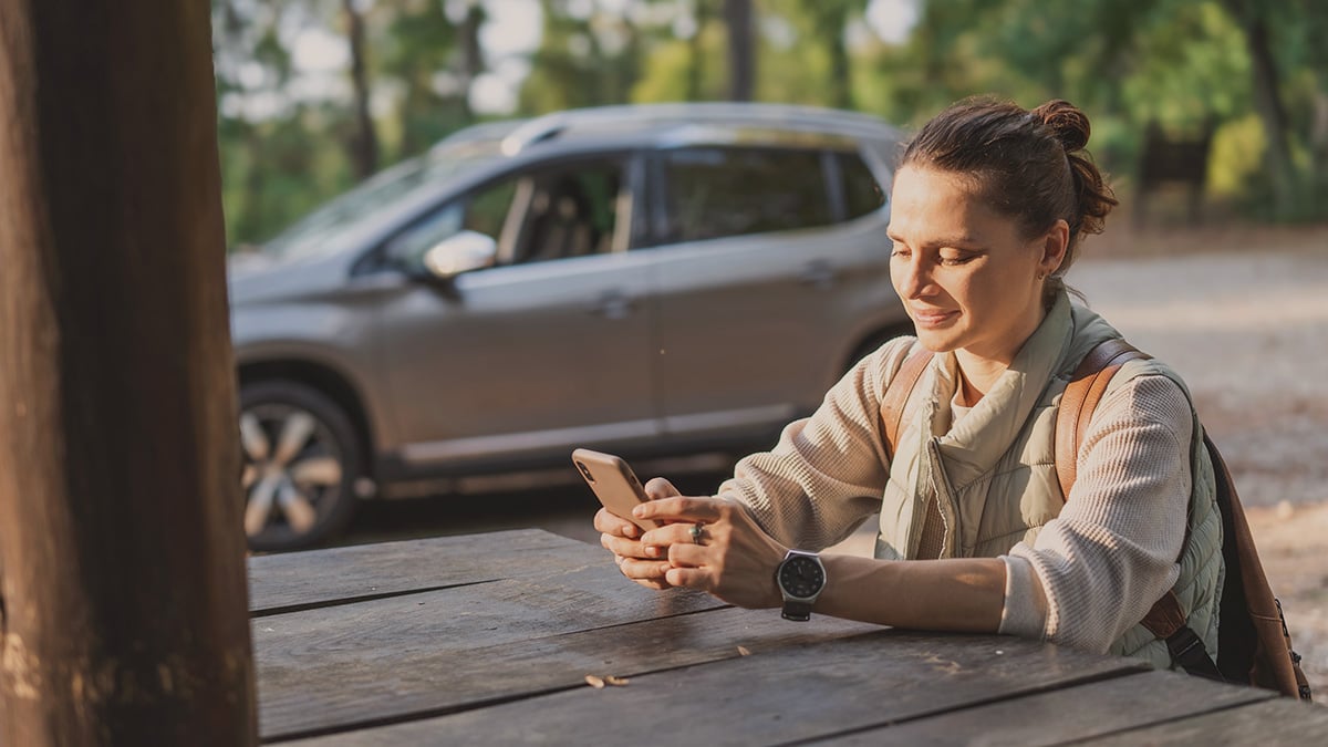 Mladá žena pozerá do mobilu na odpočívadle. Za ňou v pozadí je jej odparkované auto.