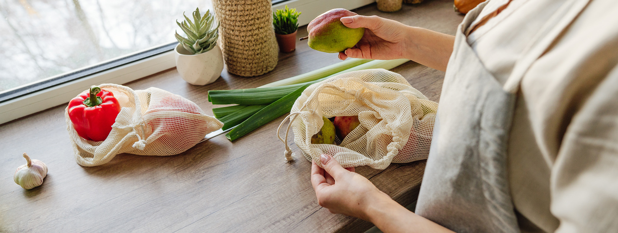 Na drevenej kuchynskej doske pri okne je položený cesnak, vedľa neho dve červené papriky vo vrecúšku a pór. V popredí vidieť ženskú postavu v krémovej zástere a tričku. V ľavej ruke drží priotvorené vrecúško, z ktorého vidieť časť hrušiek. V pravej ruke drží citrón.V popredí