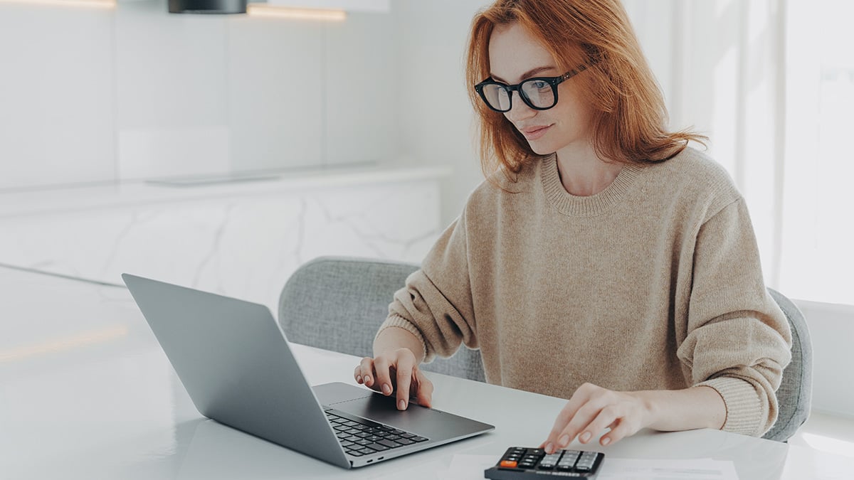 Mladá žena si udržuje prehľad o svojej finančnej situácii. Pozerá sa do notebooku a prepočítava si financie na kalkulačke. 