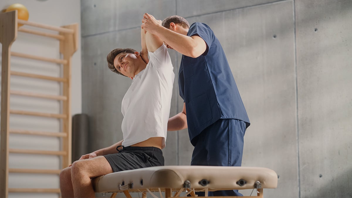 Mladý muž sedí na rehabilitácií, lekár mu pomáha natiahnuť si ruku za chrbát.