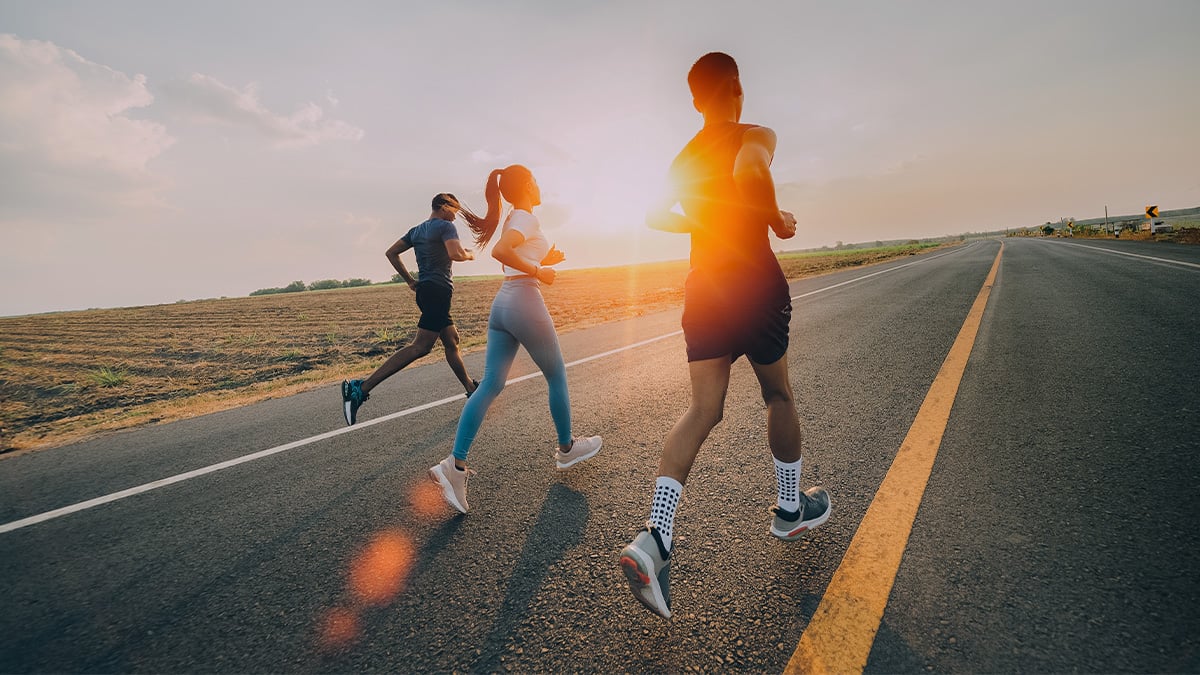 Skupina mladých ľudí v športovom oblečení beží po ceste. Slnko zapadá.