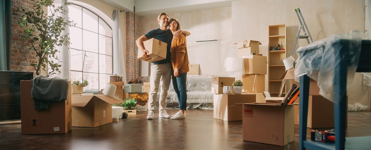 Pár sťahujúci sa do nového bývania. Muž drží krabicu a šťastne objíma manželku.