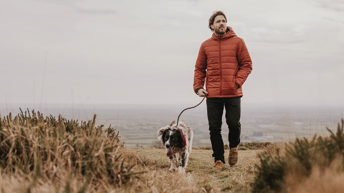 Mladý muž v oranžovej bunde, ktorý je na prechádzke so svojím psíkom v zimnom období. Kráčajú do kopca.