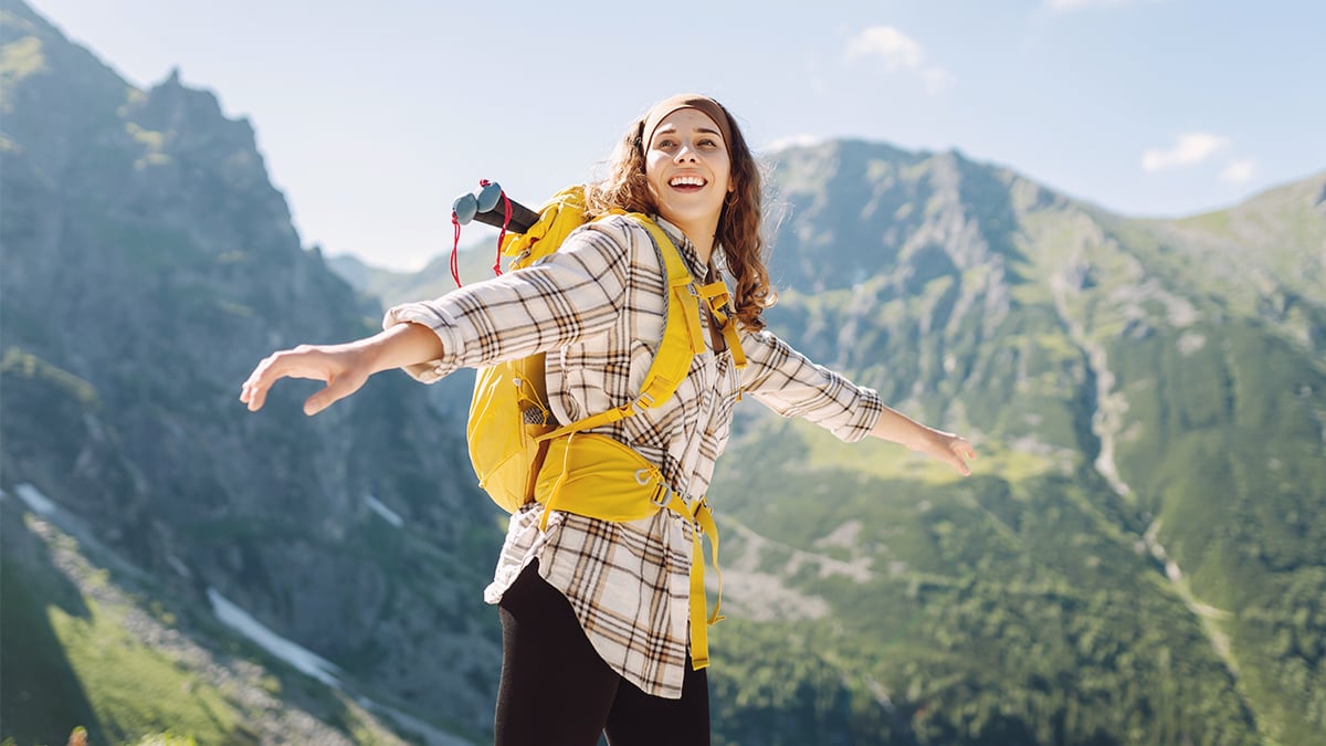 Mladá turistka, ktorá je na vrchole hôr. Má na sebe veľký žltý turistický ruksak. Za ženou je nádherná scenéria hôr..