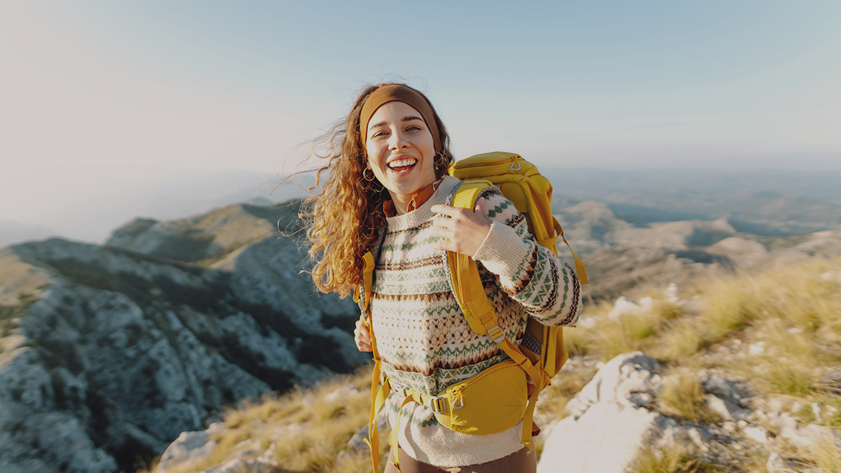 Mladá vysmiata turistka s rozviatymi kučeravými hnedými vlasmi na vrchole hôr. Má na sebe veľký žltý turistický ruksak. Za ňou je nádherná panoráma hôr.