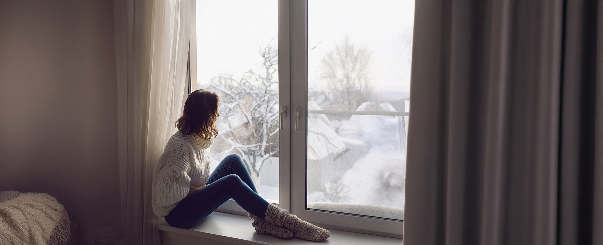 Žena v bielom svetri sedí na parapete okna, pozerá von, kde je zima a sneh.