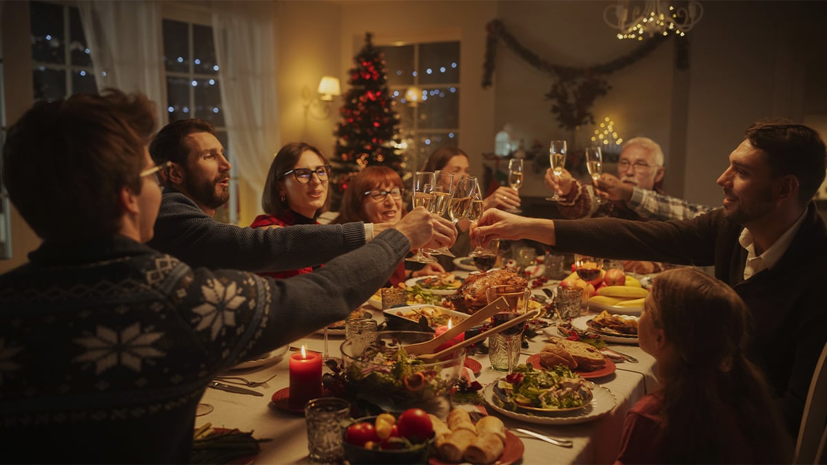 Rodina pri štedrej večeri pripíjajúca si šampanským. V rohu je vianočný stromček a na stole množstvo jedla.