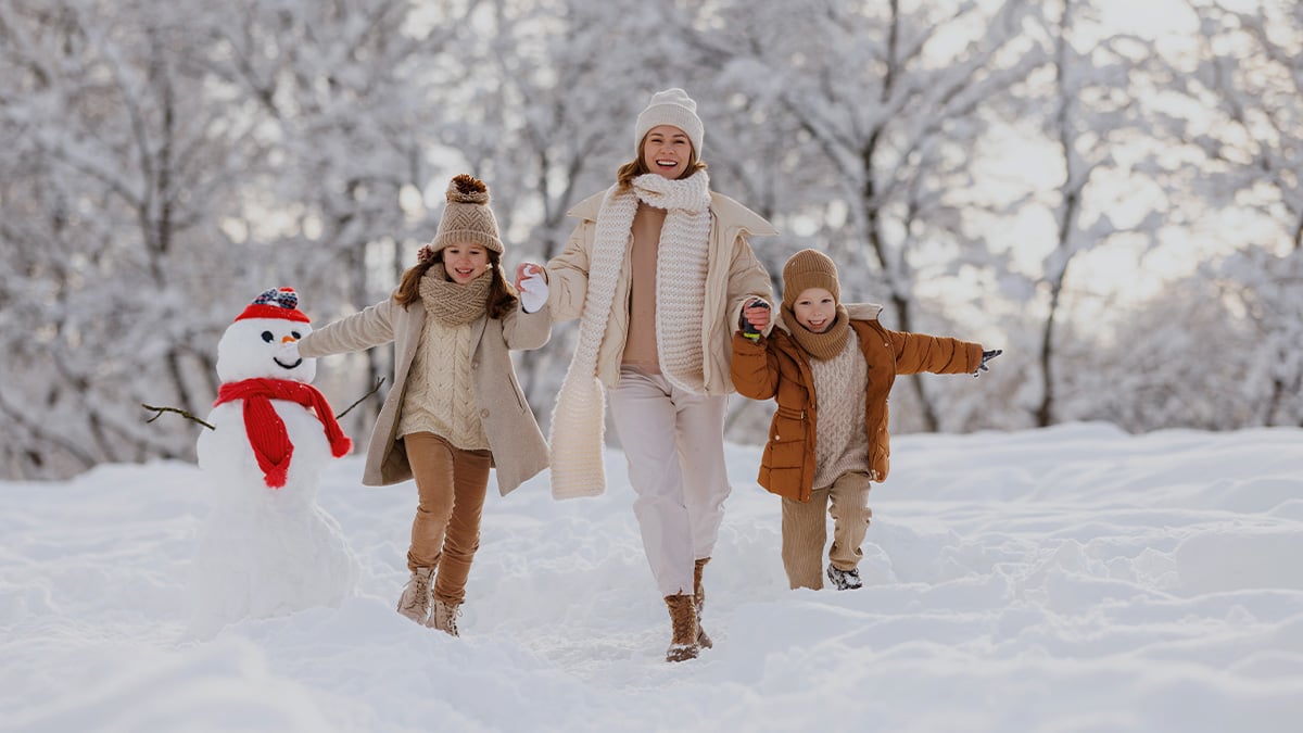 Mama s deťmi na zimnej prechádzke v prírode. Zem je pokrytá množstvom snehu, vedľa rodiny stojí snehuliak.