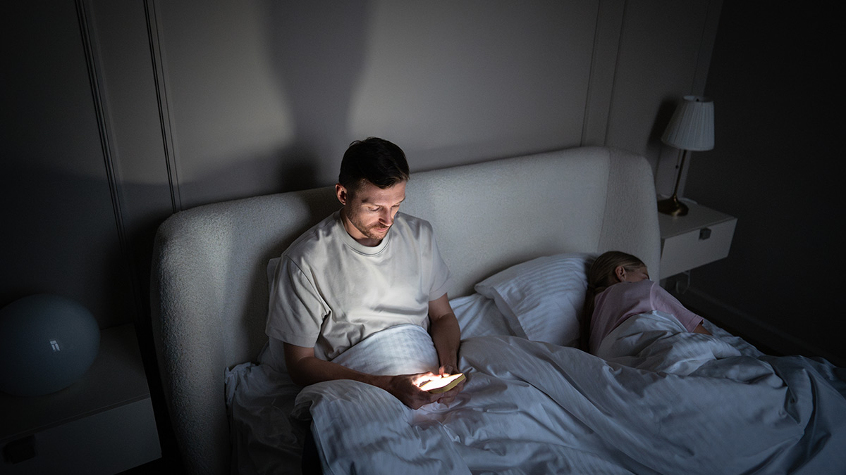 Muž v posteli pred spaním scrolluje na mobile. Hlavu má predklonenú dopredu. Po spánku môže pociťovať bolesť krčnej chrbtice. Obrazovka mobilu mu svieti do tváre.
