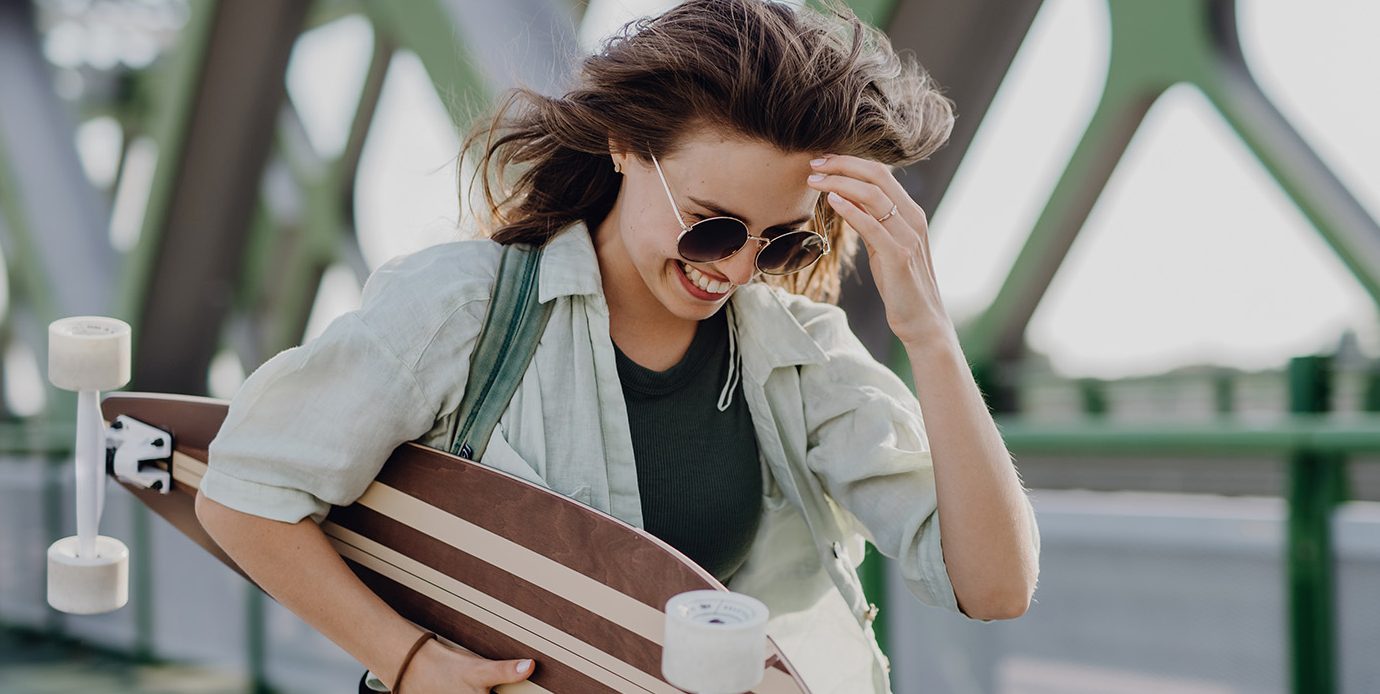 Žena nesie skateboard v rukách a usmieva sa.