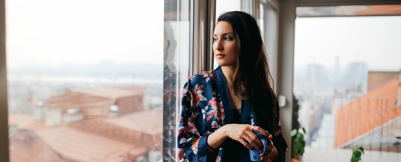 Žena v župane stoji pri presklenenom okne a pozerá sa von ,popri tom ako drží šálku.