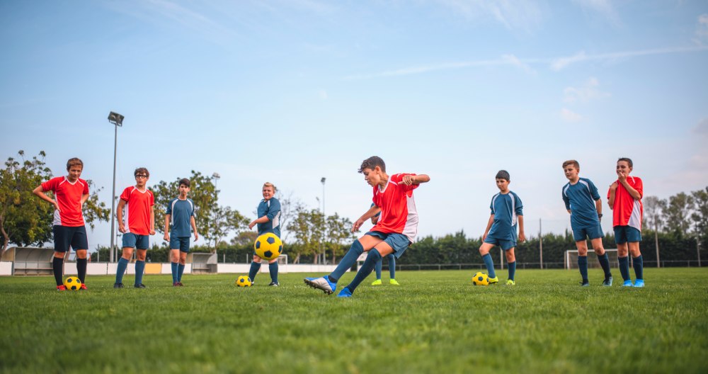Mladý člen futbalového družstva kope do lopty a ostatní futbalisti stoja v pozadí a sledujú ho.