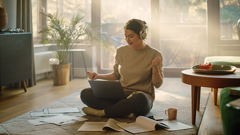 Mladá žena ponorená do práce sediaca v tureckom sede v obývačke na koberci. Na nohách má položený notebook, okolo seba má papierové brožúry, na ušiach má slúchadlá a tvári sa šťastne.