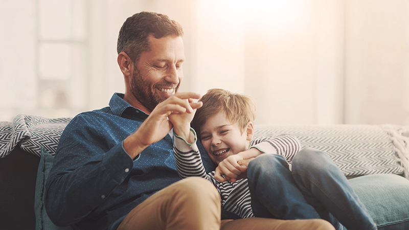 Šťastný otec s malým 8- ročným synom sedia vedľa seba na gauči v obývačke, hrajú sa a smejú. Spoza nich vidieť presvitať slnko, ktoré pohlcuje interiér.