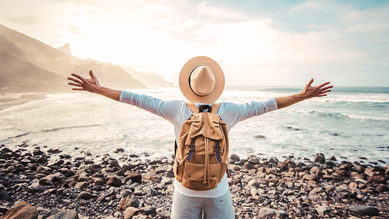 Muž s ruksakom a klobúkom stojí na kamienkovej pláži otočený chrbtom k objektívu. Muž má slobodne rozpäté ruky do strán a pozerá sa na more a skaly.