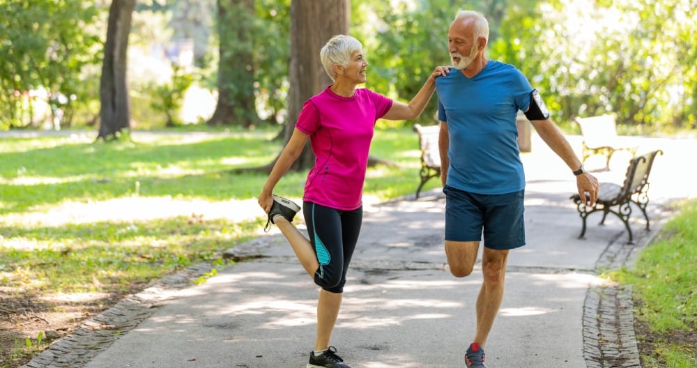 Dve staršie osoby si rozcvičujú nohy v parku.