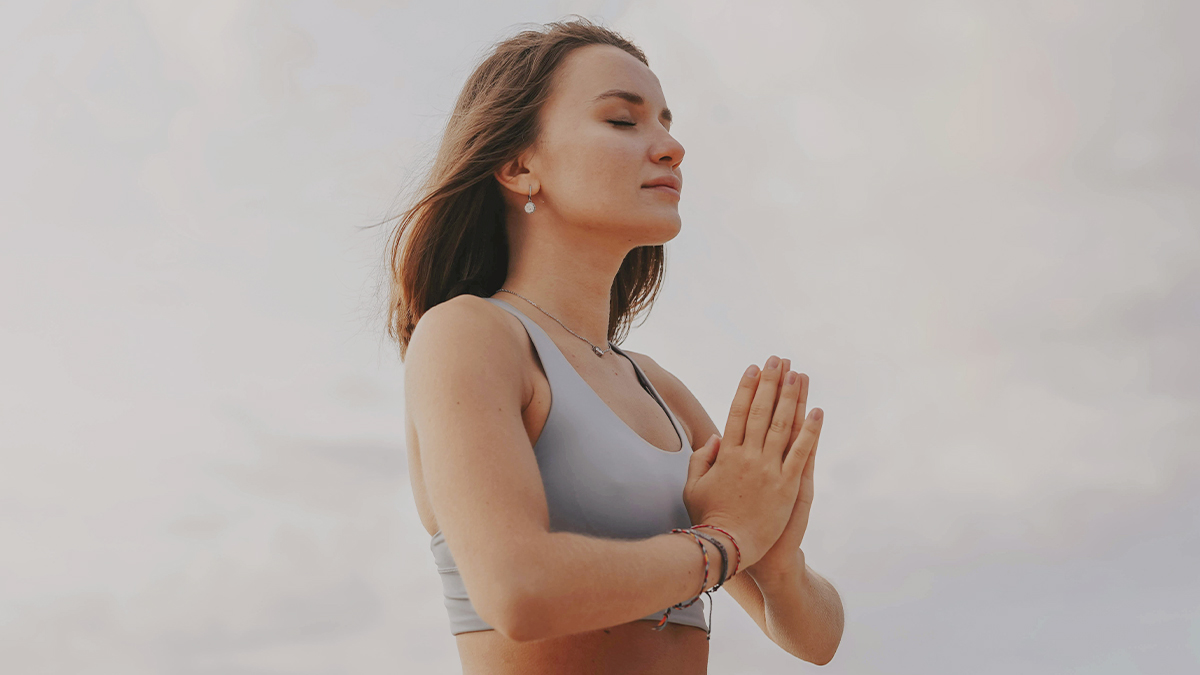 Mladšia žena v póze pri meditácii, v športovom oblečení, ruky má spojené v úrovni hrude a oči zavreté. 