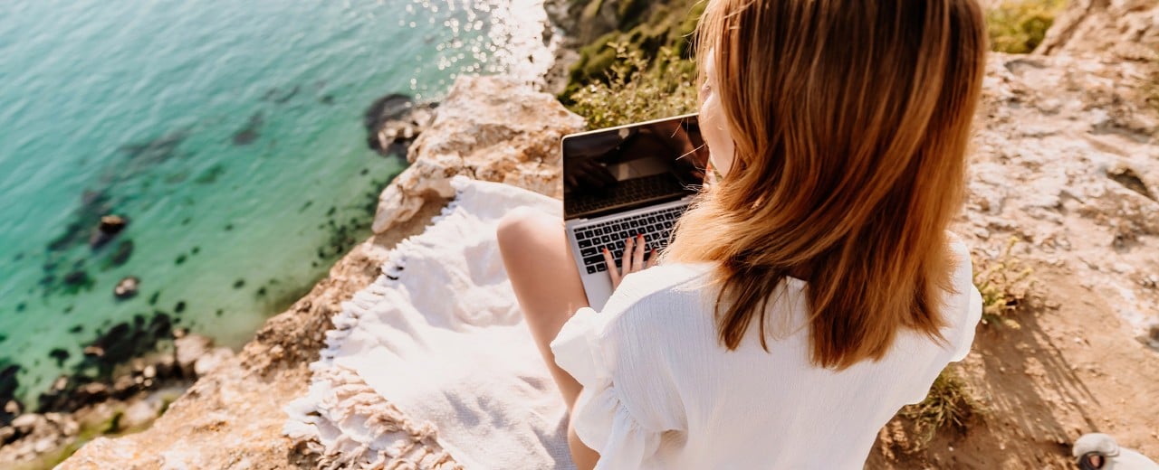 Žena v bielych letných šatách sedí na deke na vyššom útese pri mori. Na kolenách má položený notebook, na ktorom pracuje, ale pohľadom sa pozerá na vodu.  