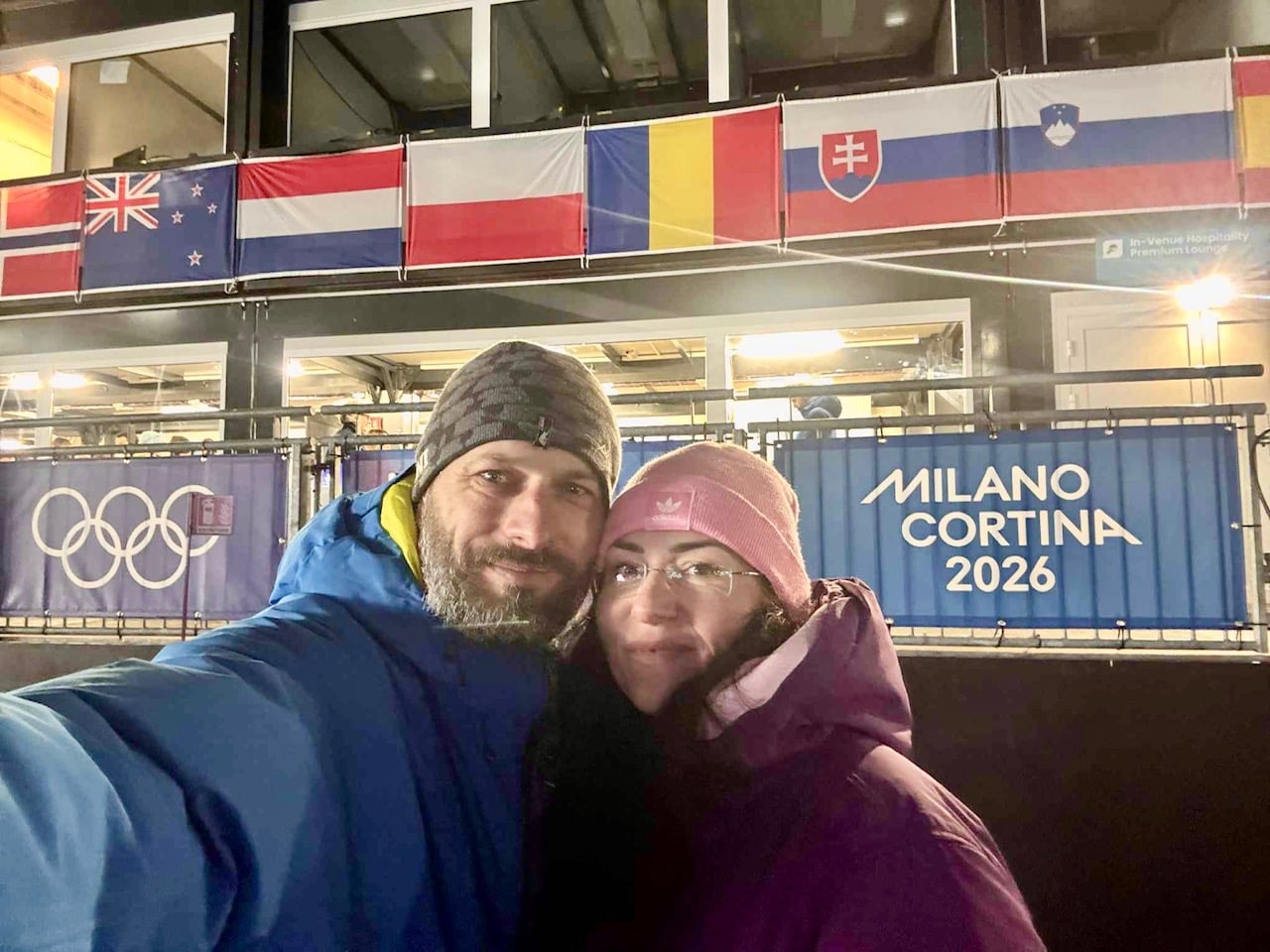 Fanúšikovia odfotení pred nápisom Miláno Cortina 2026 a vlajkami.