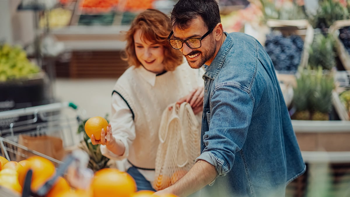 Muž so ženou vyberajú ovocie v potravinách, muž má na sebe modrú rifľovú bundu a okuliare