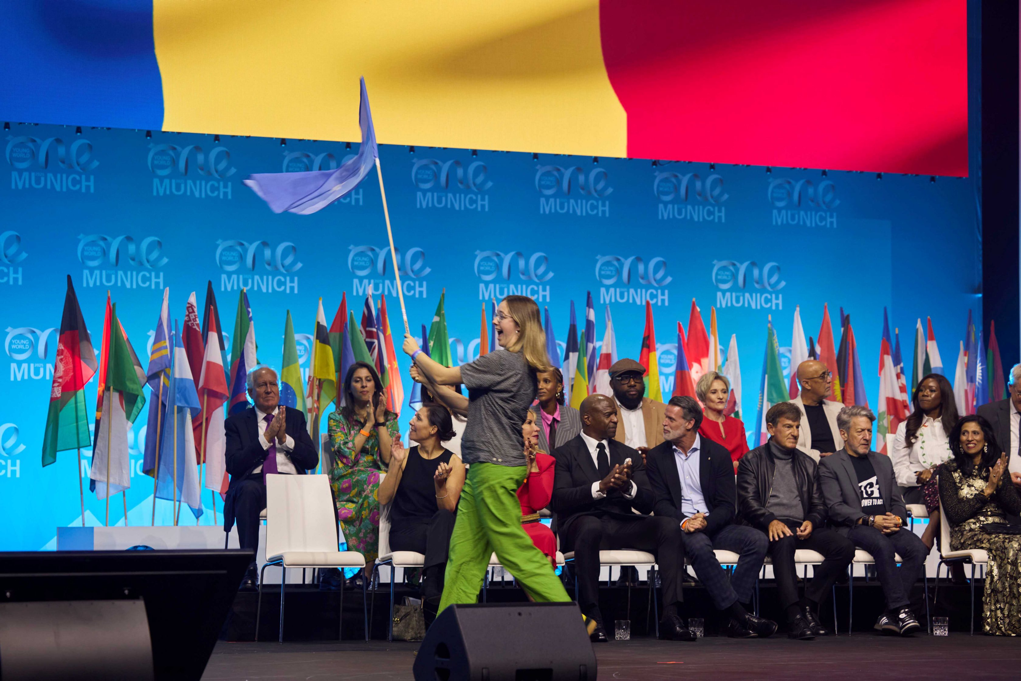Delegátka na pódiu One Young World 2025 v Mníchove máva vlajkou pred radom svetových lídrov a medzinárodných zástav.
