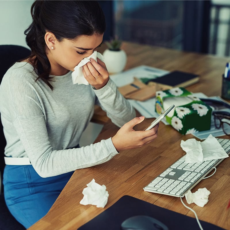 žena za stolom pozerajúca sa na mobil, utiera si vreckovkou nos