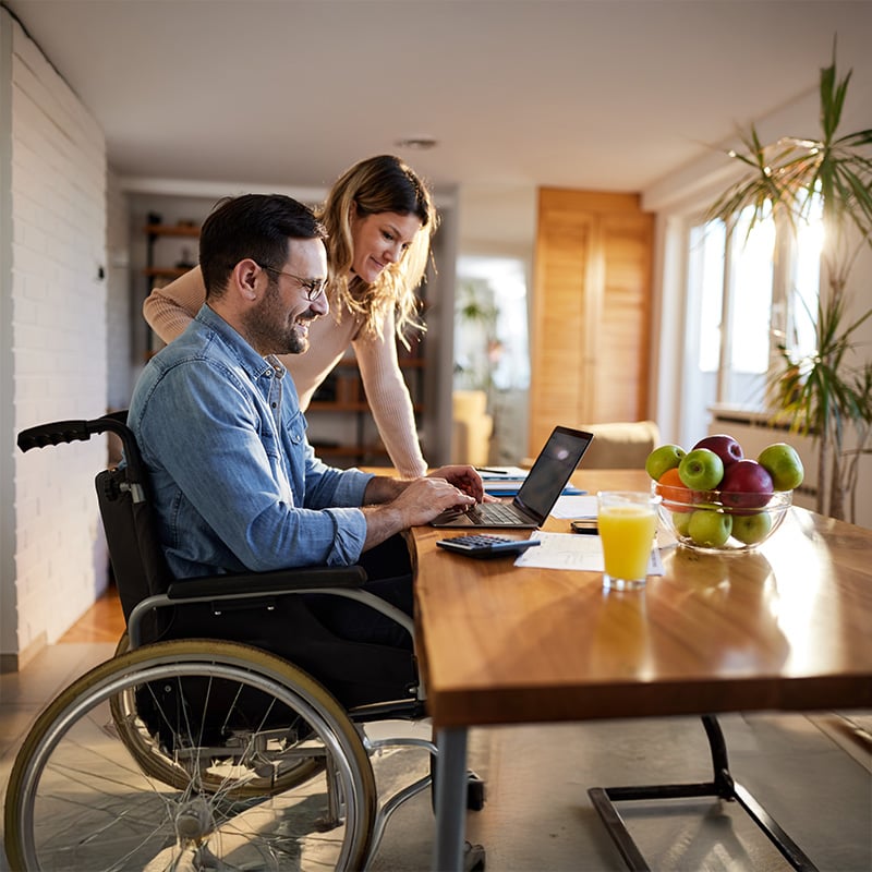 mlady muz na invalidnom voziku, zena stoji pri nom a pozeraju do pocitaca