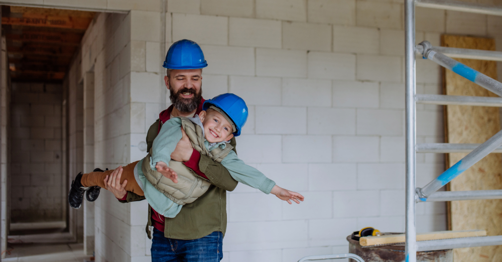 Otec drží svojho malého syna na rukách. Obaja majú na hlavách stavebné prilby pretože sa nachádzajú v stavebnom prostredí.