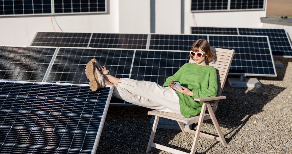 Žena sedí na stoličke vo vonkajšom prostredí a má vyložené nohy na solárnom paneli.
